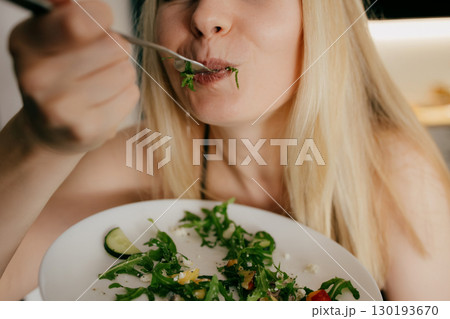 Blond woman savoring fresh salad with fork from white plate, relishing nutritious meal in her kitchen, surrounded by vibrant vegetables and wholesome ingredients. Blond woman enjoying fresh salad Blond woman savoring fresh salad with fork from white plate, relishing nutritious meal in her kitchen, surrounded by vibrant vegetables and wholesome ingredients. Blond woman enjoying fresh salad 130193670