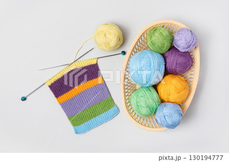 Set for the hobby of knitting warm clothes or toys. Yellow, green, blue, orange and lilac colors knitting balls in basket with knitting needles and knitted scarf on gray background. Concept needlework 130194777