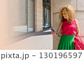 Coquettish s young female model in fashionable elegant bright multicolored green and red clothes looks at her phone against the background of the columns of an office building in the city 130196597