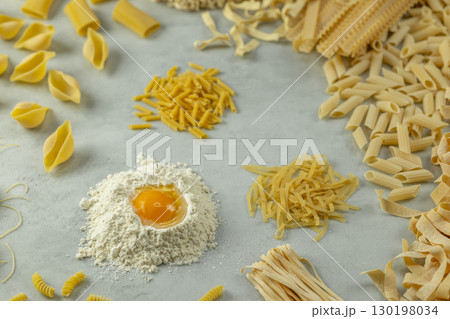 Variety of uncooked pasta with flour and egg yolk forming a circle on a gray background Variety of uncooked pasta with flour and egg yolk forming a circle on a gray background 130198034