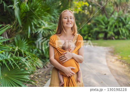 Mother holding her little baby in a yellow sling in the park. Warm and loving family moment. Babywearing, parenting, and mother-child bonding concept 130198228
