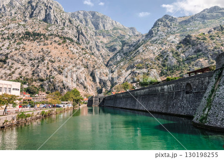 A picturesque view of the ancient city walls of Kotor, Montenegro, rising majestically alongside a tranquil canal with emerald green waters. 130198255
