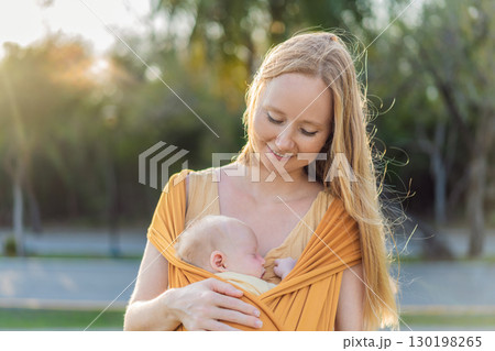Mother holding her little baby in a yellow sling in the park. Warm and loving family moment. Babywearing, parenting, and mother-child bonding concept Mother holding her little baby in a yellow sling in the park. Warm and loving family moment. Babywearing, parenting, and mother-child bonding concept 130198265