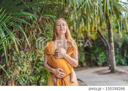 Mother holding her little baby in a yellow sling in the park. Warm and loving family moment. Babywearing, parenting, and mother-child bonding concept Mother holding her little baby in a yellow sling in the park. Warm and loving family moment. Babywearing, parenting, and mother-child bonding concept 130198266