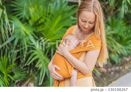 Mother holding her little baby in a yellow sling in the park. Warm and loving family moment. Babywearing, parenting, and mother-child bonding concept Mother holding her little baby in a yellow sling in the park. Warm and loving family moment. Babywearing, parenting, and mother-child bonding concept 130198267