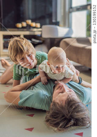 Father, son, and newborn baby lying on the floor in a cozy nursery, celebrating the arrival of their newest family member. Parenthood, bonding, and family love concept 130199152