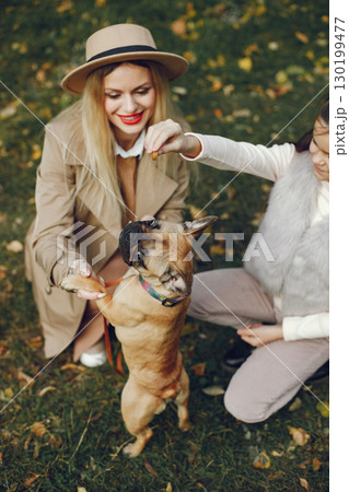 Woman, little girl and dog playing in the autumn park. Brown french bulldog with female owners spend a day at the park playing and having fun. Woman wearing beige coat and a hat. 130199477
