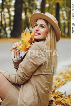 Woman posing for a photo in the autumn park. Caucasian girl has red lips. Woman wearing beige coat and a hat. 130199484