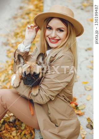 Woman and dog play and have fun in the autumn park. Brown french bulldog with female owner spend a day at the park playing and having fun. Woman wearing beige coat and a hat. 130199487