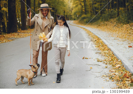 Woman, little girl and dog walking in the autumn park. Brown french bulldog with female owners spend a day at the park playing and having fun. Woman wearing beige coat and a hat. Woman, little girl and dog walking in the autumn park. Brown french bulldog with female owners spend a day at the park playing and having fun. Woman wearing beige coat and a hat. 130199489