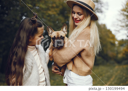 Woman, little girl and dog play and have fun in the autumn park. Brown french bulldog with female owners spend a day at the park playing and having fun. Woman wearing beige coat and a hat. 130199494