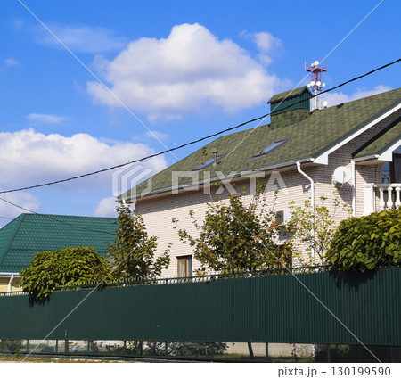Asphalt shingle. Decorative bitumen shingles on the roof of a brick house. Fence made of corrugated metal. Asphalt shingle. Decorative bitumen shingles on the roof of a brick house. Fence made of corrugated metal. 130199590