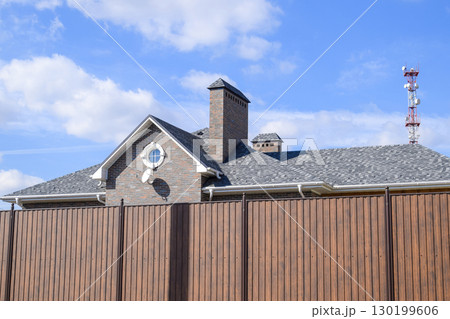 Asphalt shingle. Decorative bitumen shingles on the roof of a brick house. Fence made of corrugated metal. Asphalt shingle. Decorative bitumen shingles on the roof of a brick house. Fence made of corrugated metal. 130199606