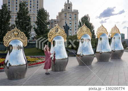 Woman in a rose dress stands beside giant reflective matryoshka sculptures with ornate golden crowns at Taras Shevchenko Embankment in Moscow 130200774