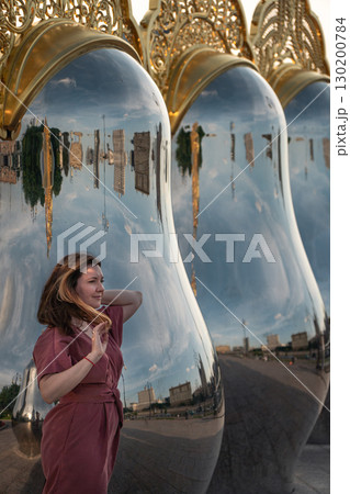 Woman in a rose dress poses by giant reflective matryoshka sculptures with ornate golden tops on Taras Shevchenko Embankment in Moscow 130200784