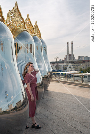 Woman in a rose dress poses by giant reflective matryoshka sculptures with ornate golden tops on Taras Shevchenko Embankment in Moscow 130200785