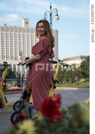 Smiling woman in a rose dress standing with an electric scooter on Taras Shevchenko Embankment, Moscow, with the White House in the background Smiling woman in a rose dress standing with an electric scooter on Taras Shevchenko Embankment, Moscow, with the White House in the background 130200790
