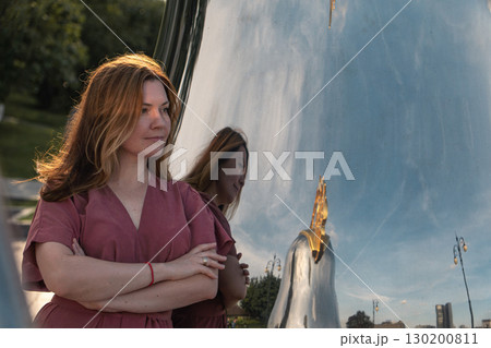 Woman in rose dress stands with arms crossed by mirrored matryoshka sculpture, evening sunlight highlighting her hair Woman in rose dress stands with arms crossed by mirrored matryoshka sculpture, evening sunlight highlighting her hair 130200811