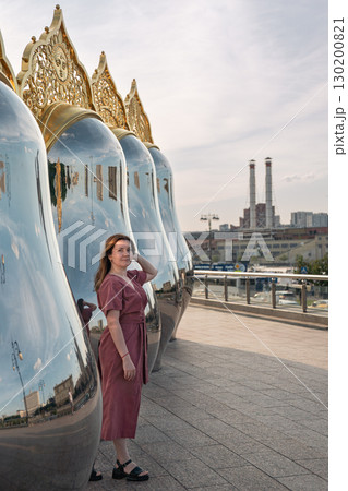 Woman in a rose dress poses by giant reflective matryoshka sculptures with ornate golden tops on Taras Shevchenko Embankment in Moscow Woman in a rose dress poses by giant reflective matryoshka sculptures with ornate golden tops on Taras Shevchenko Embankment in Moscow 130200821