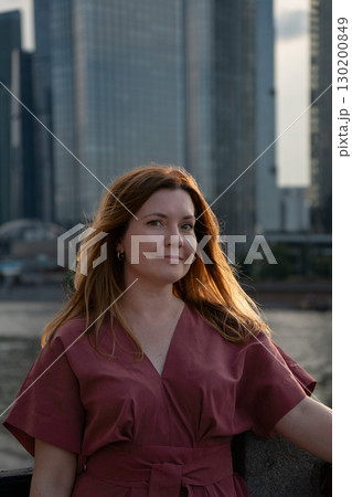 Portrait of a woman in a red dress with loose hair, softly lit by evening sunlight, standing against a background of modern skyscrapers 130200849