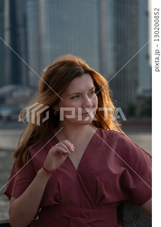Woman in a red dress with loose hair looks thoughtfully to the side, standing outdoors with blurred modern skyscrapers in the background 130200852