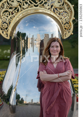 Woman in a rose dress leans against a giant reflective matryoshka sculpture with ornate golden crown on Taras Shevchenko Embankment in Moscow 130200863