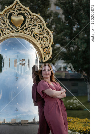 Woman in a rose dress leans against a giant reflective matryoshka sculpture with ornate golden crown on Taras Shevchenko Embankment in Moscow Woman in a rose dress leans against a giant reflective matryoshka sculpture with ornate golden crown on Taras Shevchenko Embankment in Moscow 130200873