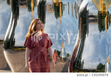 Woman in a rose dress walking among mirrored matryoshka sculptures with golden crowns on Taras Shevchenko Embankment in Moscow 130200878