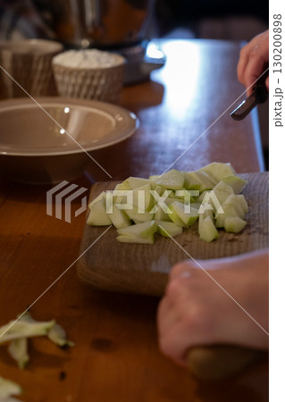 Freshly chopped green apple pieces are transferred from a wooden cutting board into a bowl, with a cup of flour visible in the background 130200898
