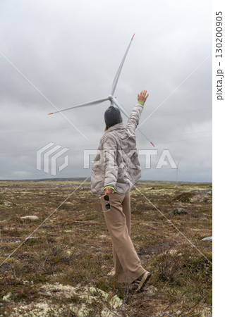 Person in a gray jacket and beanie reaches up towards a wind turbine in the tundra of Teriberka, Russia Person in a gray jacket and beanie reaches up towards a wind turbine in the tundra of Teriberka, Russia 130200905