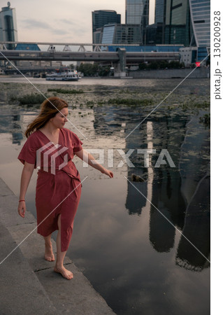 Woman in a burgundy dress walks barefoot by the Moscow River,balancing with arms outstretched against the backdrop of water and skyscraper reflections Woman in a burgundy dress walks barefoot by the Moscow River,balancing with arms outstretched against the backdrop of water and skyscraper reflections 130200928