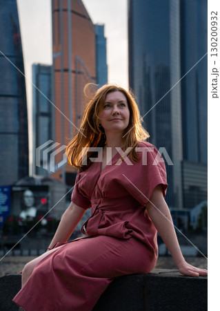 Woman in a red dress sits by the river with skyscrapers of Moscow City in the background, softly lit by sunset Woman in a red dress sits by the river with skyscrapers of Moscow City in the background, softly lit by sunset 130200932