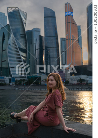 Woman in a red dress sits by the river at sunset with Moscow City skyscrapers in the background 130200933