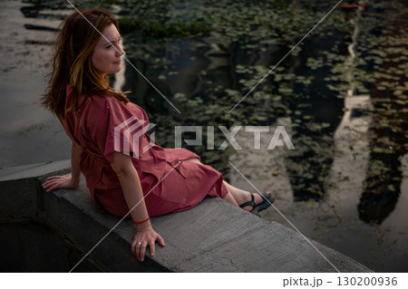 Woman in a burgundy dress sits on a stone embankment, gazing thoughtfully at the river surface with evening reflections Woman in a burgundy dress sits on a stone embankment, gazing thoughtfully at the river surface with evening reflections 130200936
