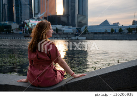 Woman in a burgundy dress sits on the Moscow River embankment at sunset, with skyscrapers of Moscow City in the background 130200939