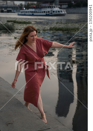 Woman in a burgundy dress walks barefoot by the Moscow River,balancing with arms outstretched against the backdrop of water and skyscraper reflections Woman in a burgundy dress walks barefoot by the Moscow River,balancing with arms outstretched against the backdrop of water and skyscraper reflections 130200941