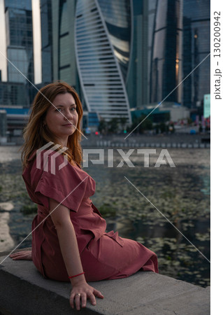Woman in a burgundy dress sits on the embankment with skyscrapers of Moscow City in the background 130200942