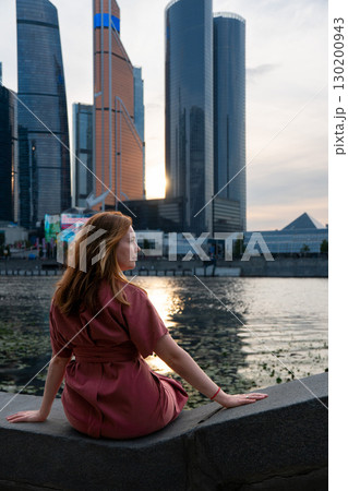 Woman in a burgundy dress sits on the Moscow River embankment at sunset, with skyscrapers of Moscow City in the background Woman in a burgundy dress sits on the Moscow River embankment at sunset, with skyscrapers of Moscow City in the background 130200943
