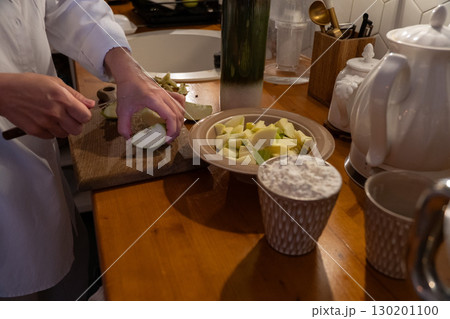 A person slices green apples on a wooden cutting board next to a bowl of chopped apples and a cup of flour on the kitchen table A person slices green apples on a wooden cutting board next to a bowl of chopped apples and a cup of flour on the kitchen table 130201100