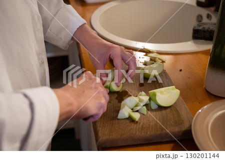A person slices green apples on a wooden cutting board next to a bowl of chopped apples and a cup of flour on the kitchen table A person slices green apples on a wooden cutting board next to a bowl of chopped apples and a cup of flour on the kitchen table 130201144