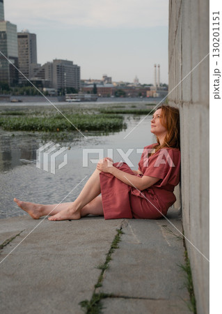 Woman in a burgundy dress sits barefoot by the Moscow River embankment, leaning against a concrete wall with skyscrapers in the background Woman in a burgundy dress sits barefoot by the Moscow River embankment, leaning against a concrete wall with skyscrapers in the background 130201151