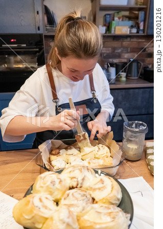 Woman spreading tasty cream onto baked cinnamon rolls on table in kitchen 130201216
