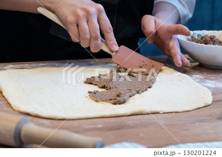 A woman spreads filling on dough with a spatula. A wooden table holds flour A woman spreads filling on dough with a spatula. A wooden table holds flour 130201224