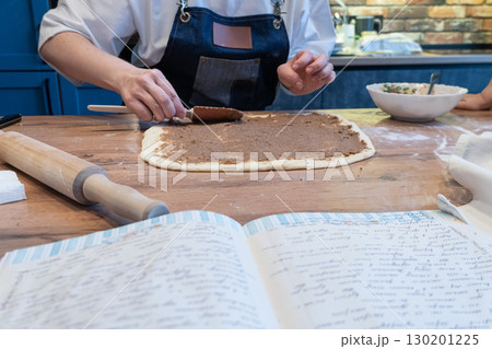 A woman spreads filling on dough with a spatula. An open cookbook with a handwritten recipe and a rolling pin are on the wooden table 130201225