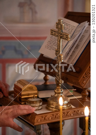 Orthodox cross standing on a carved wooden altar next to a golden box, marble container, and an open liturgical book, with candles burning in front 130201321