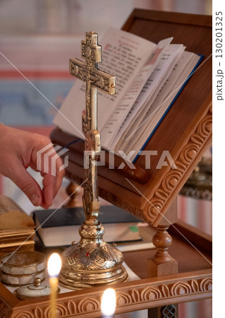 Golden Orthodox cross on a wooden altar surrounded by lit candles and sacred items, with a hand reaching toward it Golden Orthodox cross on a wooden altar surrounded by lit candles and sacred items, with a hand reaching toward it 130201325