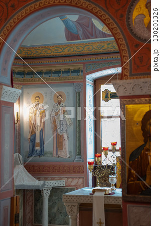 Interior of an Eastern Orthodox church with vivid iconography, frescoes of saints, and a decorated altar with red candle holders and a gold cross 130201326