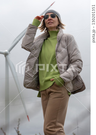 Woman in a beige puffer jacket and gray beanie adjusts her sunglasses with a wind turbine softly blurred in the background 130201351