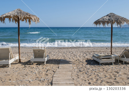 Simple beach scene on Crete - sunbeds and palm leaf umbrellas by the pebbles and sea Simple beach scene on Crete - sunbeds and palm leaf umbrellas by the pebbles and sea 130203336
