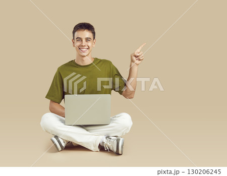 Happy teenage student boy with laptop computer pointing finger away on beige background 130206245